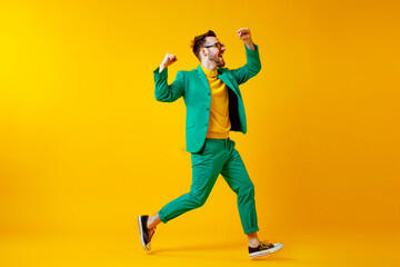 Fototapeta na wymiar Young man celebrating success. Happy funny joyful excited guy in stylish green party suit and cool glasses raising fist up and dancing isolated on bright yellow background. Full length shot, side view