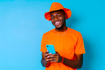 Full body young smiling happy african american man 20s in orange shirt hat use hold mobile cell phone chatting isolated on plain pastel light blue background studio portrait. People lifestyle concept
