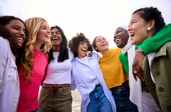 Large group of multiracial young only women standing embracing in circle laughing looking each other outdoor. Gen z female community friends people hugging together having fun enjoying leisure time