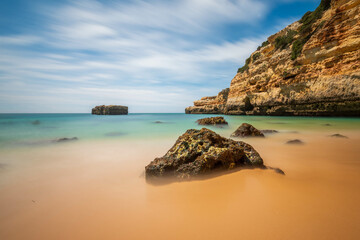 Dreamy long exposure of a secluded beach with golden sand and turquoise water, framed by dramatic...