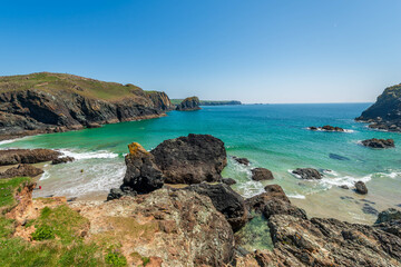 The rugged coastline of Cornwall, England, reveals a hidden cove with turquoise waters, a sandy beach, and dramatic rock formations under a clear blue sky, a breathtaking display of natural beauty.