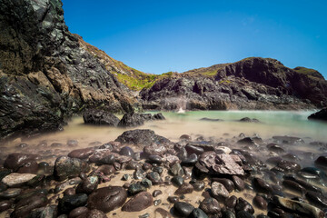 A rocky beach in Cornwall, England, with dark, dramatic rocks leading to a secluded cove with turquoise water under a vibrant blue sky, creates a captivating scene of coastal beauty.