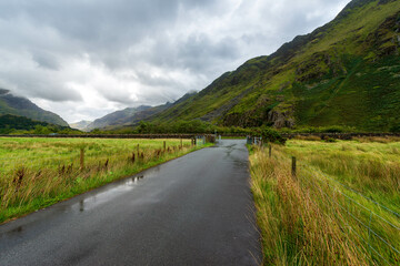A wet, winding road cuts through a verdant valley in the Scottish Highlands, with towering mountains shrouded in dramatic clouds creating a moody and atmospheric scene.