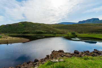 A tranquil scene of a serene lake nestled amidst rolling green hills in the Scottish Highlands, with a bright blue sky dotted with fluffy clouds reflected in the calm waters.