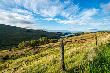 A lush green valley in the Irish countryside, with a lake nestled between rolling hills, a rustic fence in the foreground, and a dramatic sky with wispy clouds.