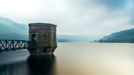alybont Reservoir's serene waters embrace a lone tower on a misty day, reflecting in the tranquil...