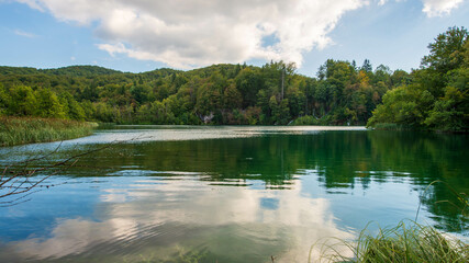 Fototapeta premium he serene waters of a lake in Plitvice Lakes National Park reflect the surrounding lush forests and cloudy sky, creating a tranquil and idyllic natural scene.