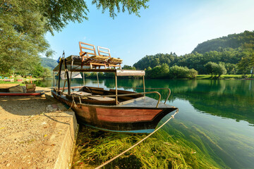 Obraz premium A traditional wooden boat rests peacefully on the serene Una River, surrounded by lush greenery and reflecting the clear blue sky, offering a tranquil escape in the heart of nature