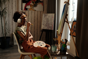 Young Black artist sitting in studio, wearing headphones and holding paintbrush while painting on...