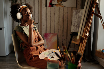 Artist sitting in cozy studio wearing headphones, holding paintbrush under chin while deep in...