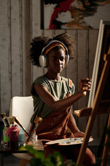 Young Black girl painting on canvas while wearing headphones in cozy studio environment. Various painting tools surrounding her work area