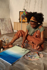 African American woman engrossed in painting serene ocean scene on canvas while sitting...