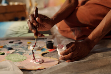Obraz premium Hands skillfully holding palette knife and mixing paints on canvas. Background reveals artistic studio ambience with various art supplies and tools