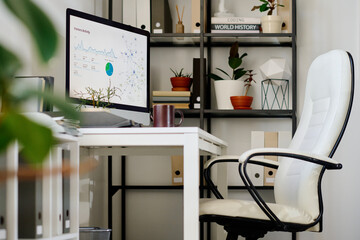 Modern white office space featuring computer monitor, shelves filled with books and plants, and comfortable chair inviting organized work atmosphere