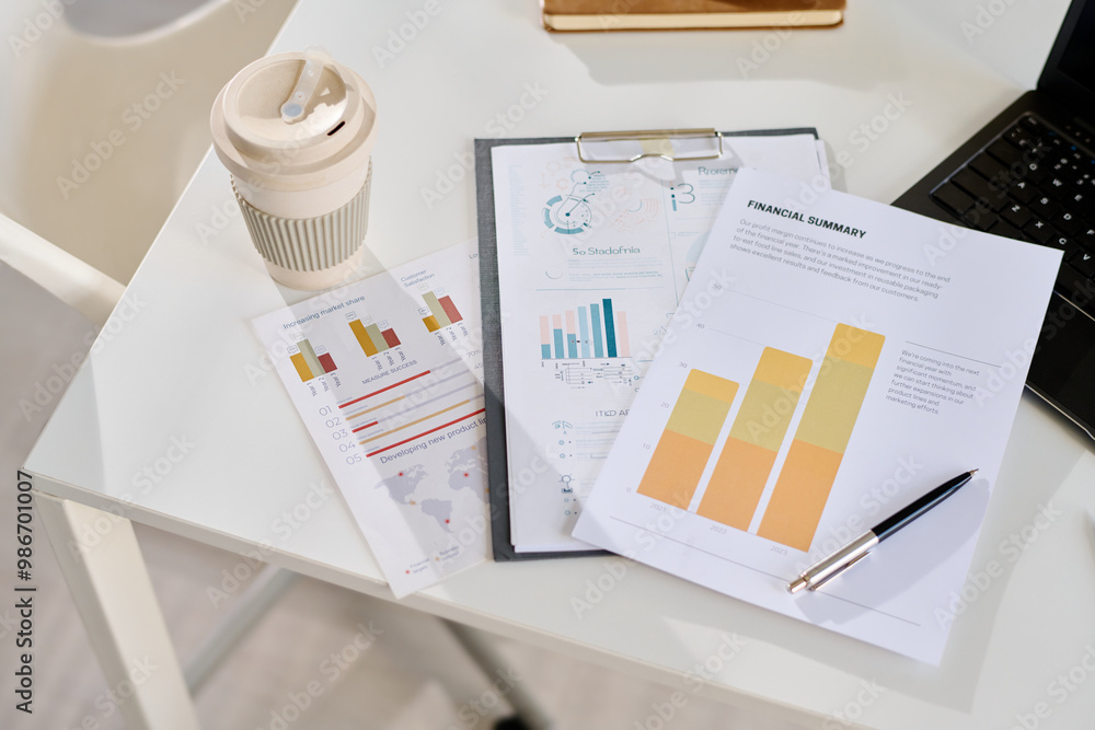 Poster financial documents with charts and graphs placed on white desk by a laptop, alongside reusable coff