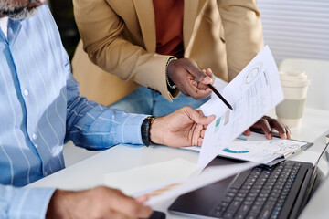Diverse team analyzing business reports in modern office with documents, coffee cup, and laptop present. Close collaboration and teamwork evident during work process