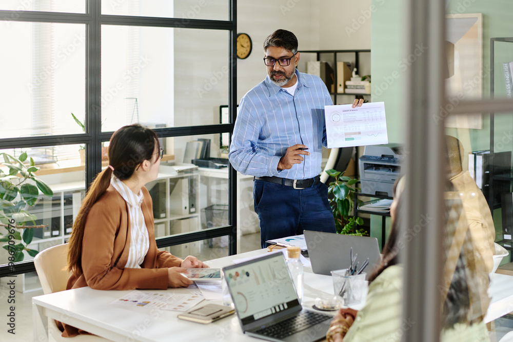 Poster Middle-aged man holding business report while presenting in modern office space. Colleagues seated at table listening attentively with laptops open for note-taking