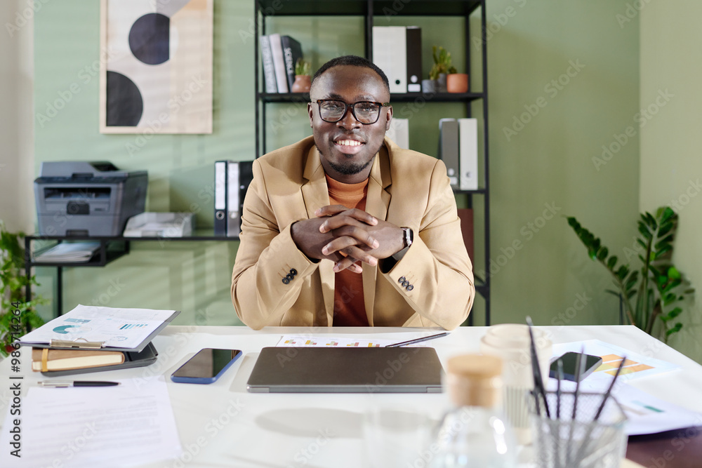 Poster portrait of smiling african american man sitting at office desk with hands clasped, surrounded by wo