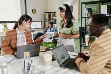 Group of diverse professionals working on project in modern office, discussing documents with laptops open on desk showing data and graphs