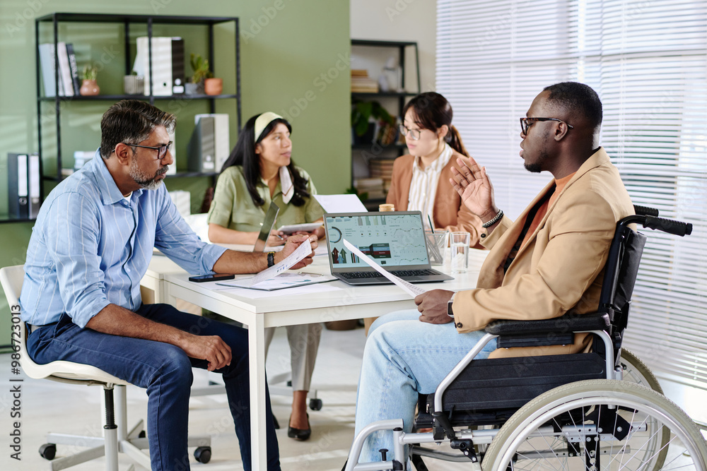 Aufkleber Group sitting around table in office discussing strategies while one man in wheelchair presenting data using laptop engaging team members in conversation