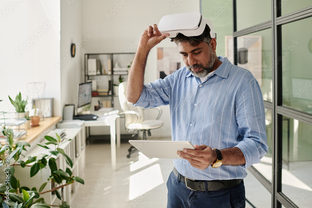 Poster Man wearing virtual reality headset and looking at tablet while standing in bright modern office with open spaces and greenery adding touch of nature