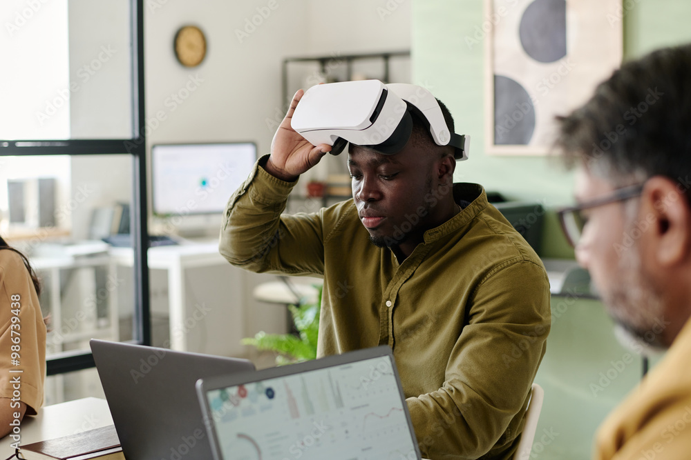 Aufkleber man engaging with virtual reality headset, sitting at desk with laptop in modern office featuring co