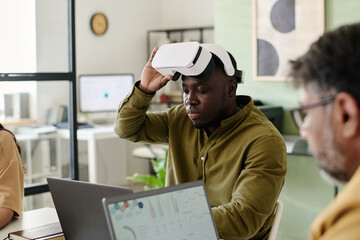 Man engaging with virtual reality headset, sitting at desk with laptop in modern office featuring coworkers and tech setup in background