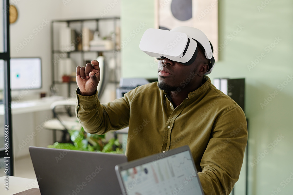 Poster Man sitting at desk wearing virtual reality headset, interacting with virtual elements. Modern office setting visible with shelves and plants creating a realistic backdrop