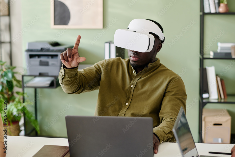 Poster Person engaging with virtual reality in an office environment with bookshelves and printer in background while seated at desk.