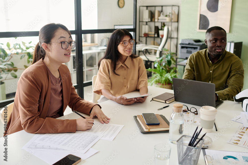 Poster asian woman in glasses leading presentation with notebook while colleagues listen attentively in mod