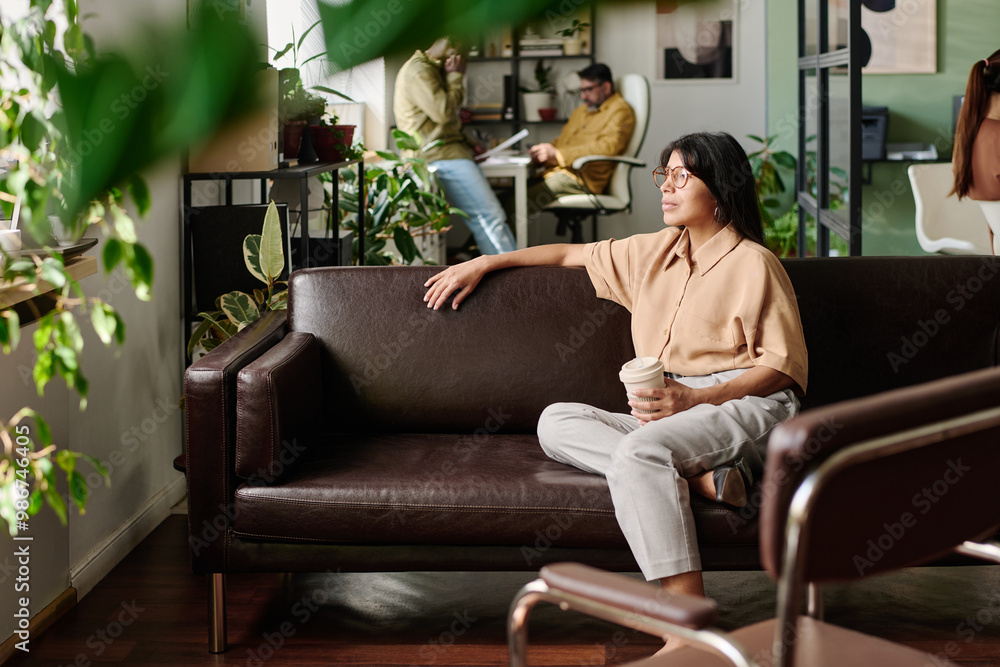 Poster office with employees engaging in tasks and relaxing on a brown sofa while holding a mug, with green
