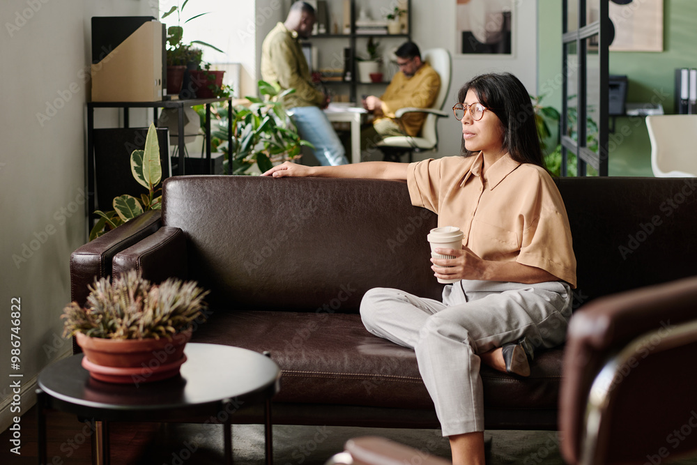Poster young woman sitting on couch holding coffee cup while coworkers work in background. modern office en