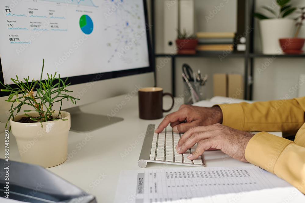 Poster Person typing on keyboard while analyzing data on computer screen and reviewing printed report. Desktop decorated with potted plants and a brown coffee mug