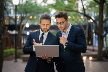 Two businessman on the street, deeply engrossed, eyes fixed on their laptop screen. Two handsome...