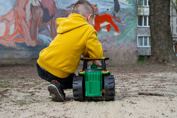 A boy in a yellow jacket is sitting on a tree stump. A boy plays with a toy car in the sand. A person is using a yellow plastic shovel to dig sand. High quality photo