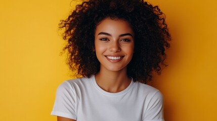 Young woman with curly hair wearing a white t-shirt, standing against a pastel yellow background, relaxed smile.