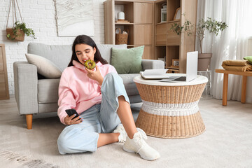 Beautiful young woman in stylish hoodie with donut and mobile phone sitting at home