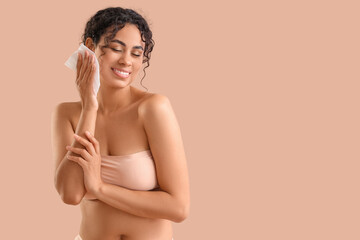 Young African-American woman removing makeup with wipe on beige background