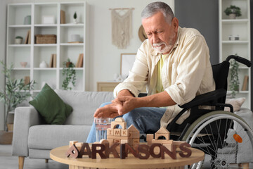 Senior man suffering from Parkinson syndrome in wheelchair taking glass of water from table at home