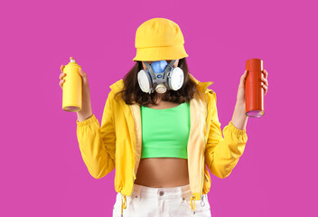 Young woman in respirator with spray paint cans on purple background