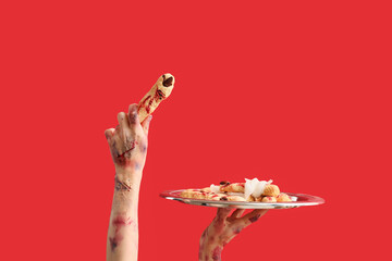 Zombie hands holding plate with cookies in shape of fingers on red background. Halloween celebration