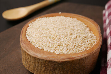A wooden bowl filled with white sesame seeds, accompanied by a spoon, set on a dark surface