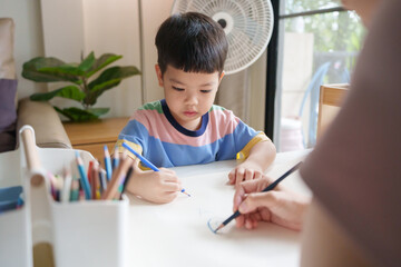 Asian young boy is drawing on a paper with a colored pencil with his mother at home.