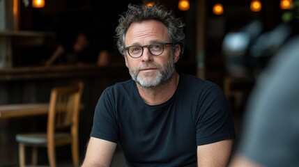 Obraz premium A thoughtful man with curly hair and glasses sits at a table in a warm cafГ© setting, illuminated by soft lighting. He appears introspective while surrounded by a relaxed atmosphere