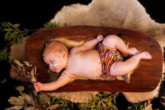 Happy Aboriginal baby boy lying on possum skin in coolamon cradle holding gum leaves