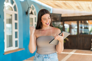 Young pretty Brazilian woman holding a tablet at outdoors celebrating a victory