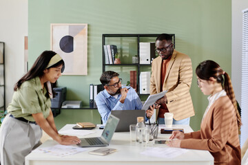 Obraz premium Office team discussing business strategy with focus on tablet Held by team member Standing. Papers and laptops are on the table while diverse team Engaged in discussion