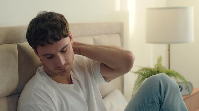 Young Caucasian man sitting on bed touching neck pain ill unhealthy fatigue massage home bedroom ache discomfort disease feeling unwell sad suffering unhappy male guy irritated face rubbing head upset