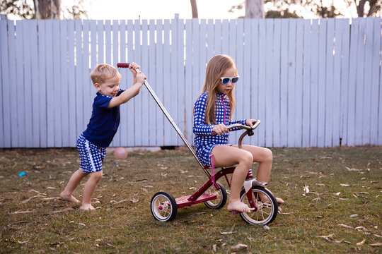 kids playing on a trike in the back yard with a girl riding and a boy pushing