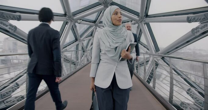 Tilt-up slow motion portrait of Middle Eastern business lady in hijab walking in stylish hall looking at city through glass walls. Business and successful people concept.